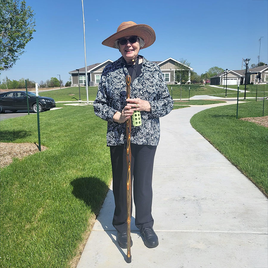 Elderly woman with cane on sidewalk, enjoying a stroll with a wooden walking stick and sun hat.