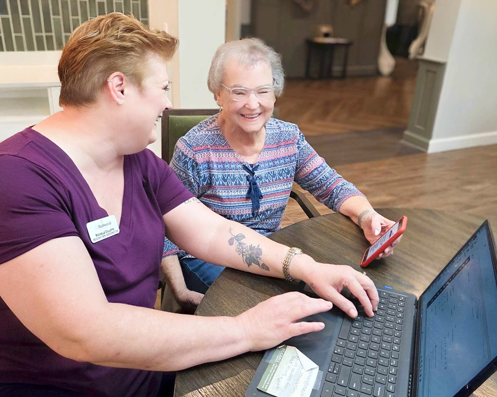 Team member helps a smiling senior resident navigate the Arrow app on her phone while seated at a round table with a laptop in a senior living community.