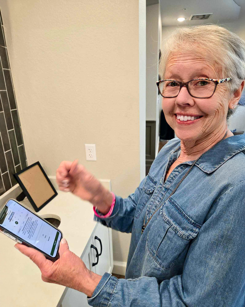 Senior resident wearing glasses and a denim shirt smiles while using the Arrow app on her phone near a modern counter in a bright senior living space.