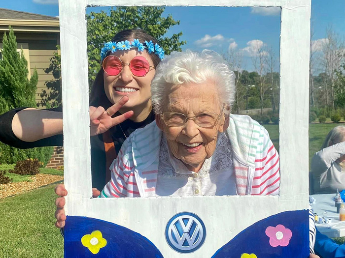 Smiling senior resident and a young woman wearing 60s-themed accessories, posing together in a VW bus cutout during an outdoor event, capturing the joy and fun of the moment.