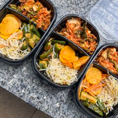 Three meal prep containers filled with a variety of vegetables, including kimchi, zucchini, yellow squash, and seasoned bean sprouts, placed on a granite countertop.