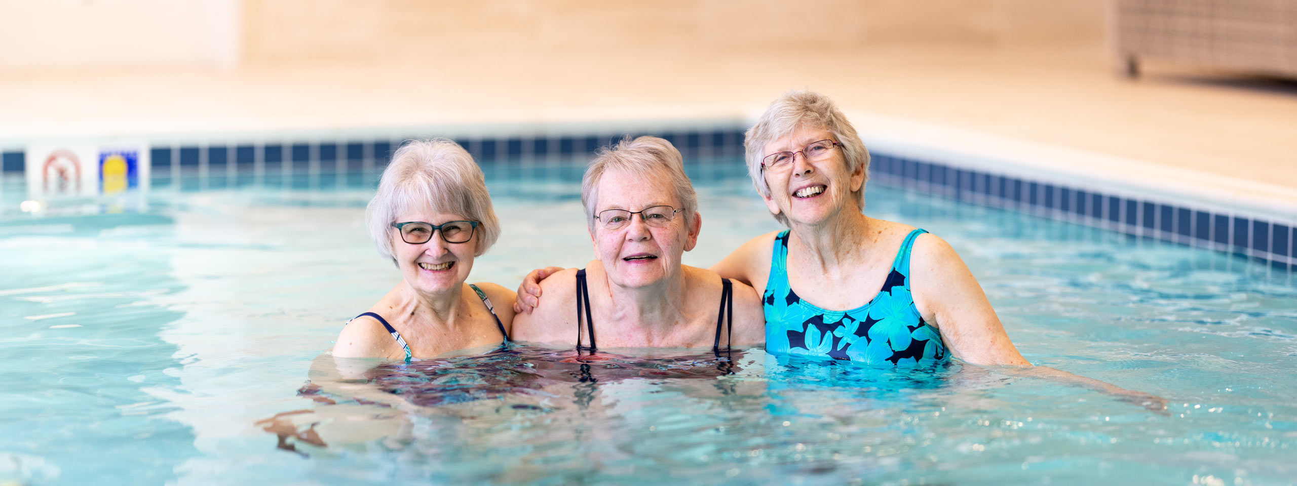Three smiling senior residents enjoying a refreshing time together in a swimming pool, showcasing active living, friendship, and the joy of fitness in a senior living community.