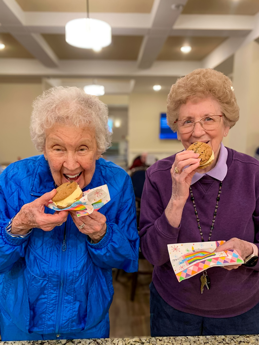 Two senior residents smiling and enjoying ice cream sandwiches in a bright and cheerful setting, holding colorful napkins, and sharing a moment of joy together.