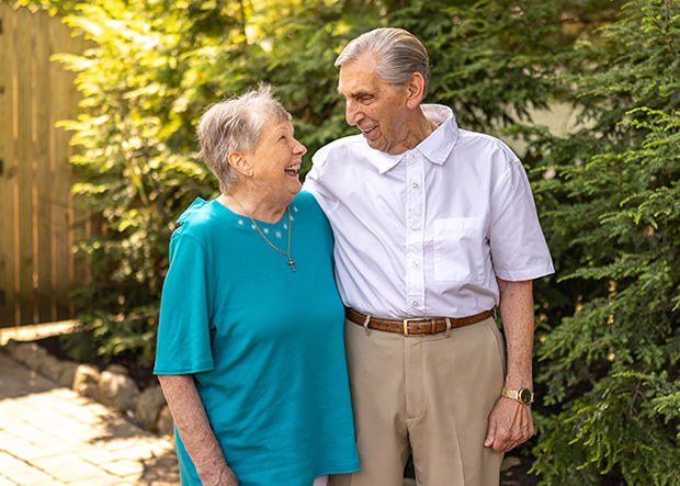 Smiling senior couple standing close together in a lush green garden, the woman wearing a teal shirt and the man in a white shirt, sharing a joyful moment in the sunlight.