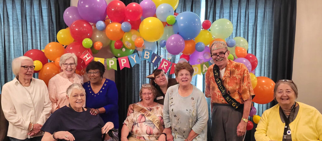 A group of senior residents celebrates a birthday in front of colorful balloons and a "Happy Birthday" banner, with the birthday king and queen seated.