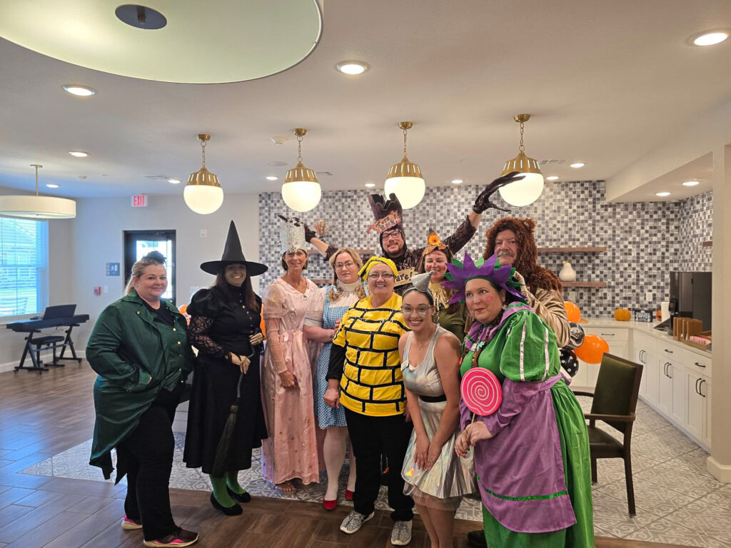 A group photo of team members dressed in fun and creative costumes, standing together in a brightly lit community area, showcasing their festive spirit.