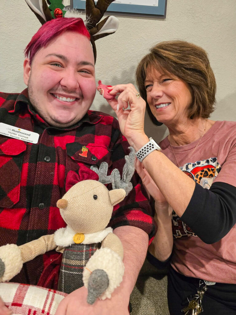 Two team members smiling together while one holds a small holiday decoration and the other displays a cheerful plush reindeer, embodying community warmth and joy.