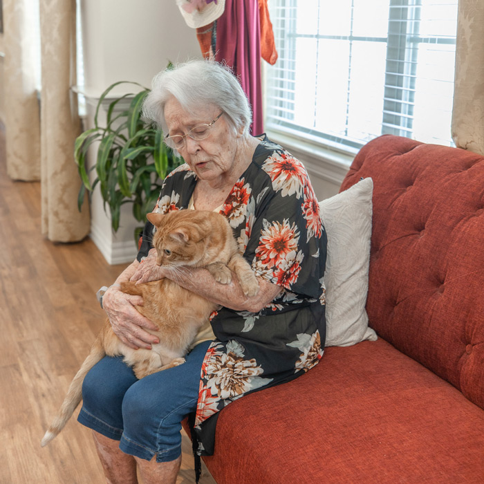 An elderly woman with short white hair and glasses is sitting on a red couch, holding a large orange cat in her lap.