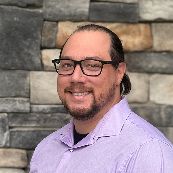 Nick Dysarz smiling man with glasses, short beard, and tied-back dark hair wears a lavender button-up shirt, posing against a stone wall background in a professional headshot.