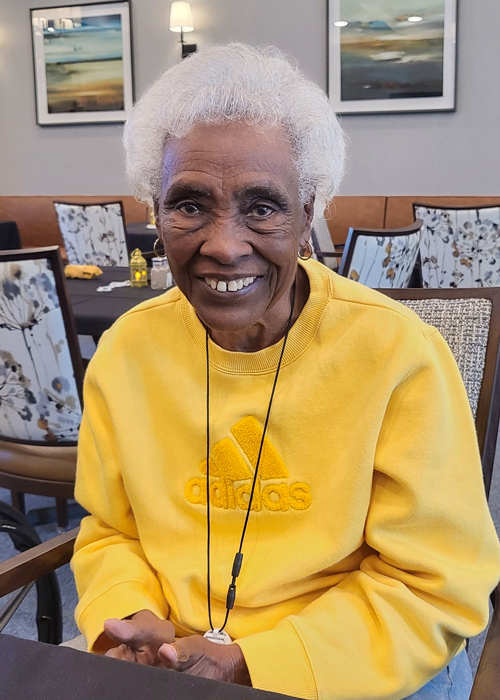 Senior resident smiles brightly while wearing a cheerful yellow Adidas sweatshirt, enjoying time in a cozy and welcoming community dining space.