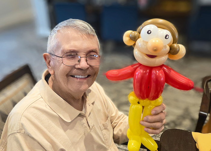 Senior resident proudly holds a colorful balloon sculpture of a cartoon figure, celebrating Food on a Stick Day with laughter and creativity.