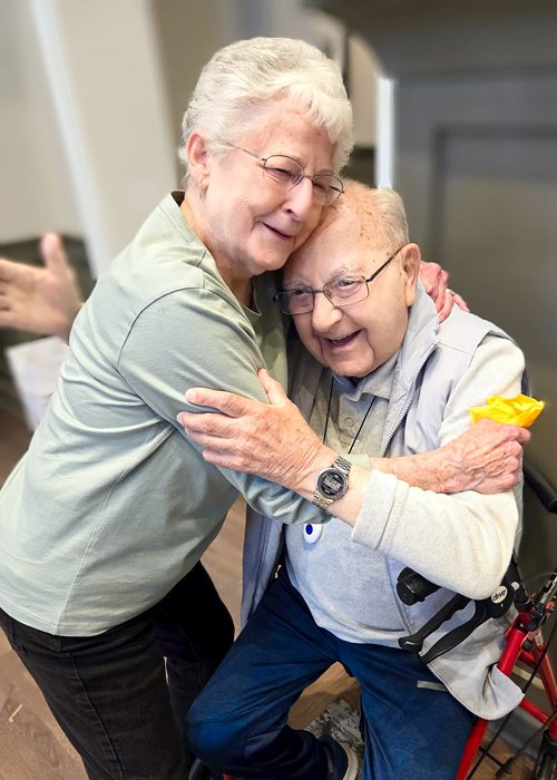 Two senior residents share a joyful embrace during a Grand Celebration event, capturing a heartfelt moment of friendship and connection.