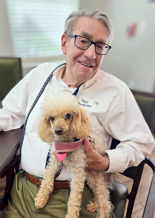 Senior resident sits with a fluffy poodle on his lap, smiling warmly during a visit with his pet in a relaxed and pet-friendly community environment.