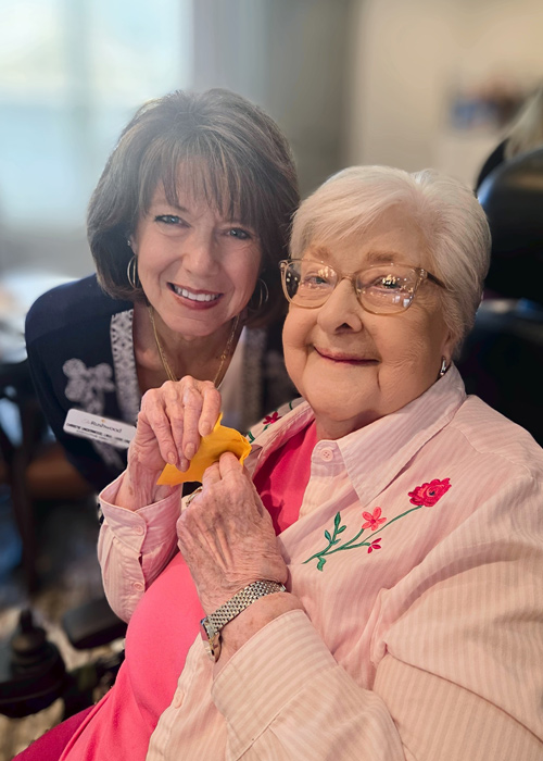 Two women smiling together during a community celebration, one seated in a wheelchair holding a yellow-shaped note