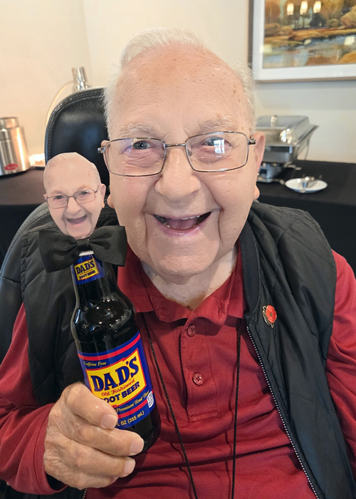 Smiling senior man holding a Dad’s Root Beer bottle decorated with a miniature cutout of his own face for Father’s Day