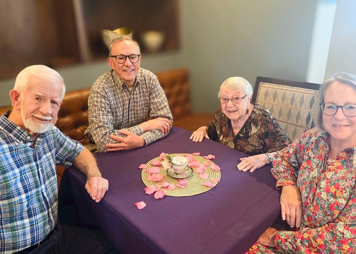 A woman in medium grey hair, glasses, and a bright floral button up shirt, another with white short hair, glasses, earrings, and a dark floral patterned shirt, and two men, both wearing plaid buttoned shirts, sit at a table covered in purple table cloth, decorated with flower petals and a floral cup and saucer set filled with sugar cubes, before sharing a meal.