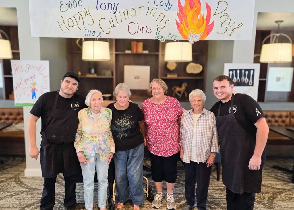 Culinary team members pose with residents under a festive banner celebrating Culinary Day, highlighting appreciation for delicious meals and joyful service.