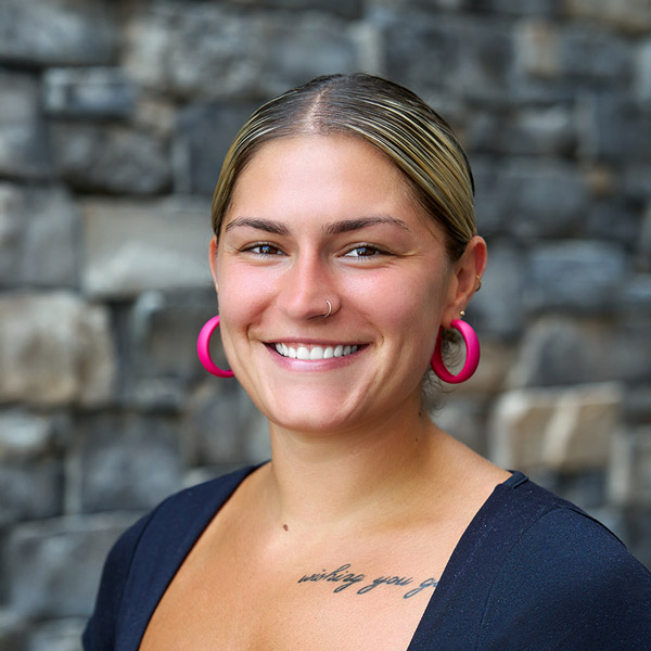 Jaylen Knight, Senior Living Coordinator at The Rushwood Senior Living, smiling in a professional headshot. She has blonde hair pulled back, wears bright pink hoop earrings and a dark top, with a script tattoo visible on her chest, standing in front of a stone wall background.