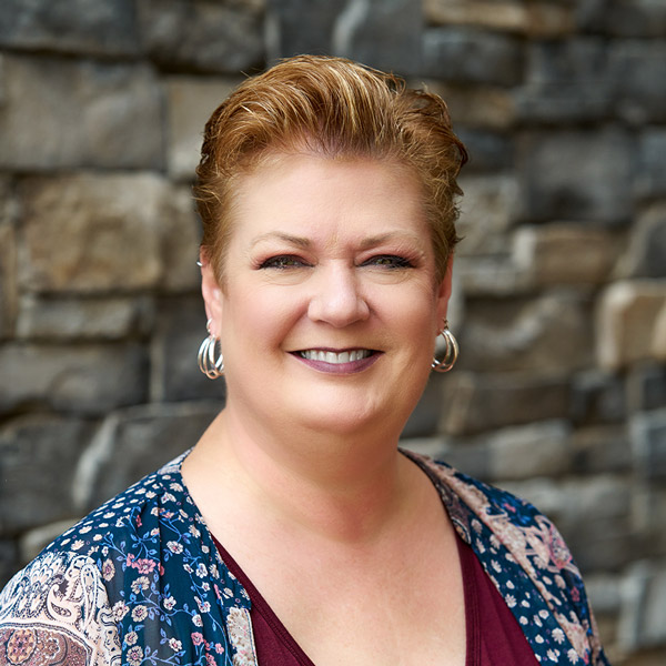 Rochelle Collette, Resident Services Director at The Rushwood Senior Living, smiling in a professional headshot. She has short, styled auburn hair, wears silver hoop earrings, a burgundy top, and a patterned floral cover-up, standing in front of a stone wall background.