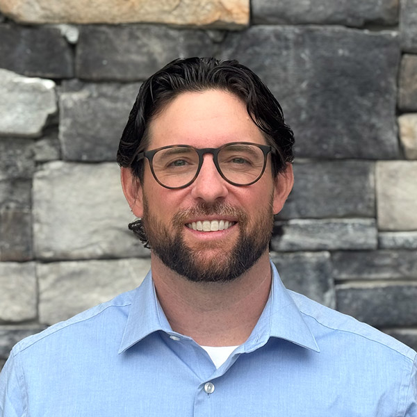 Nathan Harrison, Executive Director at The Rushwood Senior Living, smiling in a professional headshot. He has medium-length dark hair, a short beard, and wears round glasses with a light blue collared shirt, standing in front of a gray stone wall background.