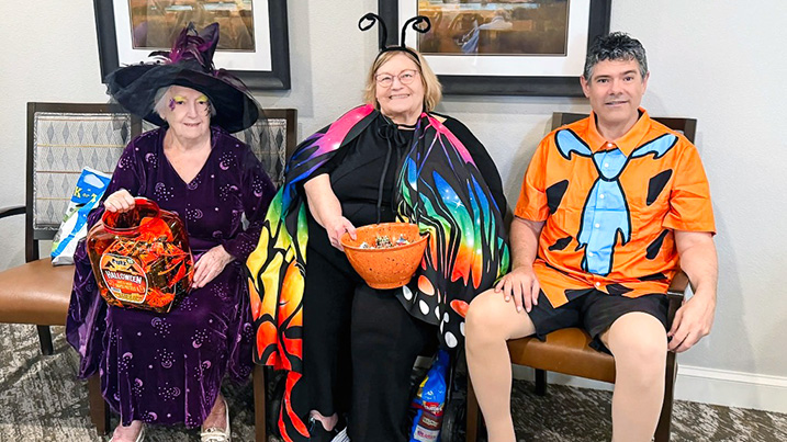 Three friends dressed in colorful costumes sit together, ready to hand out candy to trick or treaters.