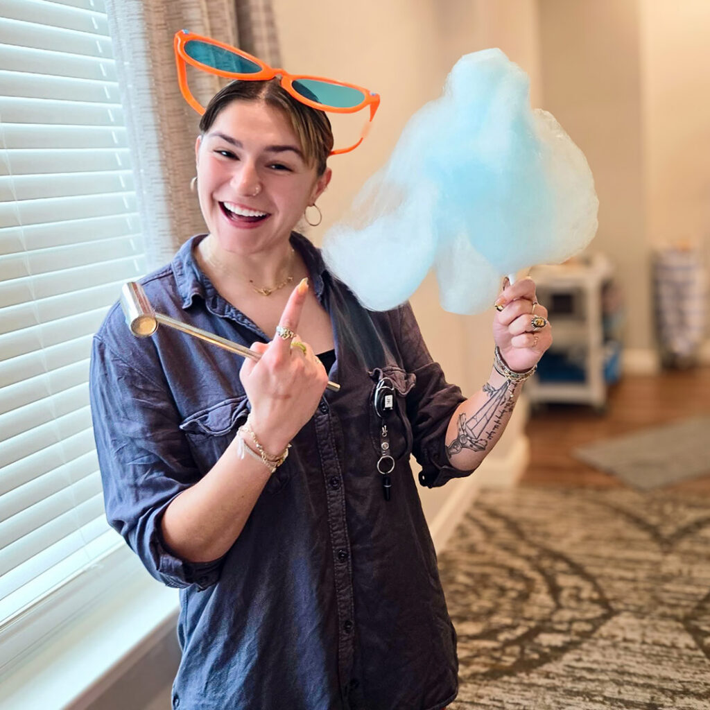 A team member at The Rushwood Senior Living smiles brightly, wearing large novelty sunglasses and holding a bright blue cotton candy.