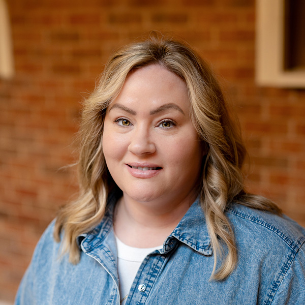 Amy Garcia, Assistant Executive Director at The Rushwood Senior Living, looking at the camera in a professional outdoor headshot with shoulder-length wavy blonde hair, a denim button-up shirt, and a softly blurred brick background.