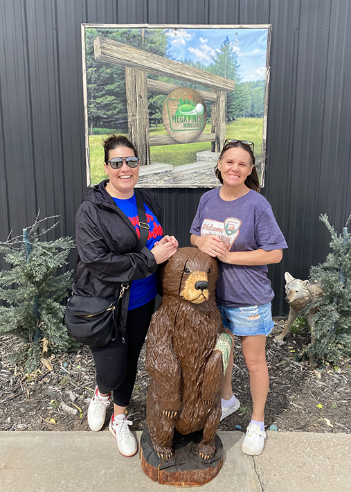 Two women smile brightly on a sunny day, resting their hands on a bear statue beside a sign for Mega Pines Mini Golf.