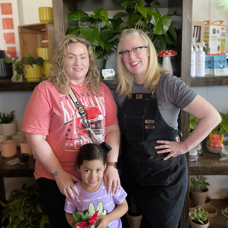 Two women and child smile brightly at a local businesses, surrounded by plants and terracotta pots.