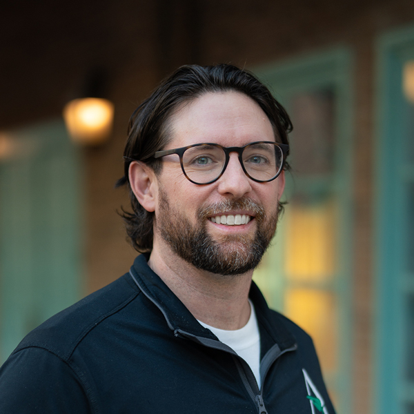 Nathan Harrison, Executive Director at The Rushwood Senior Living, smiling in a professional outdoor headshot with dark hair, glasses, a trimmed beard, and softly blurred brick and teal window details in the background.