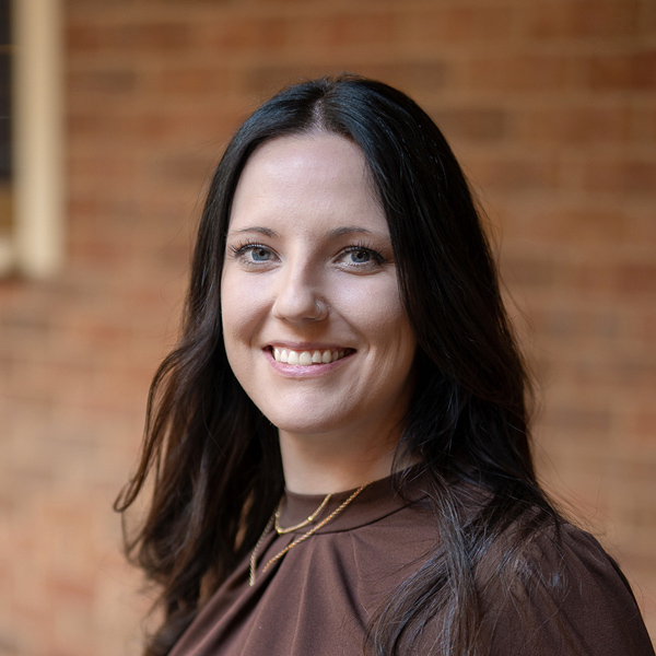 Lauren Smith, Wellness Director at The Rushwood Senior Living, smiling in a professional outdoor headshot with long dark wavy hair, a brown blouse, layered necklaces, and a softly blurred brick background.